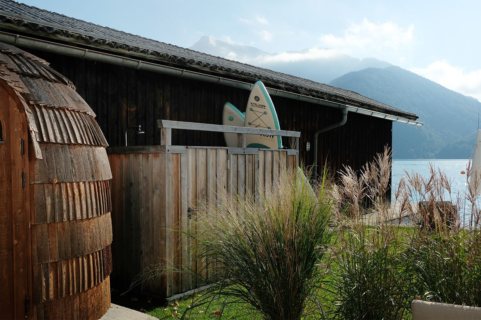 Gräser bewegen sich sachte im Wind und das Wasser glitzert; Von der Lounge geht es direkt zur großen Terrasse mit Sitzmöglichkeiten, Outdoor Kitchen, Sauna, Außendusche, SUPs und Liegewiese